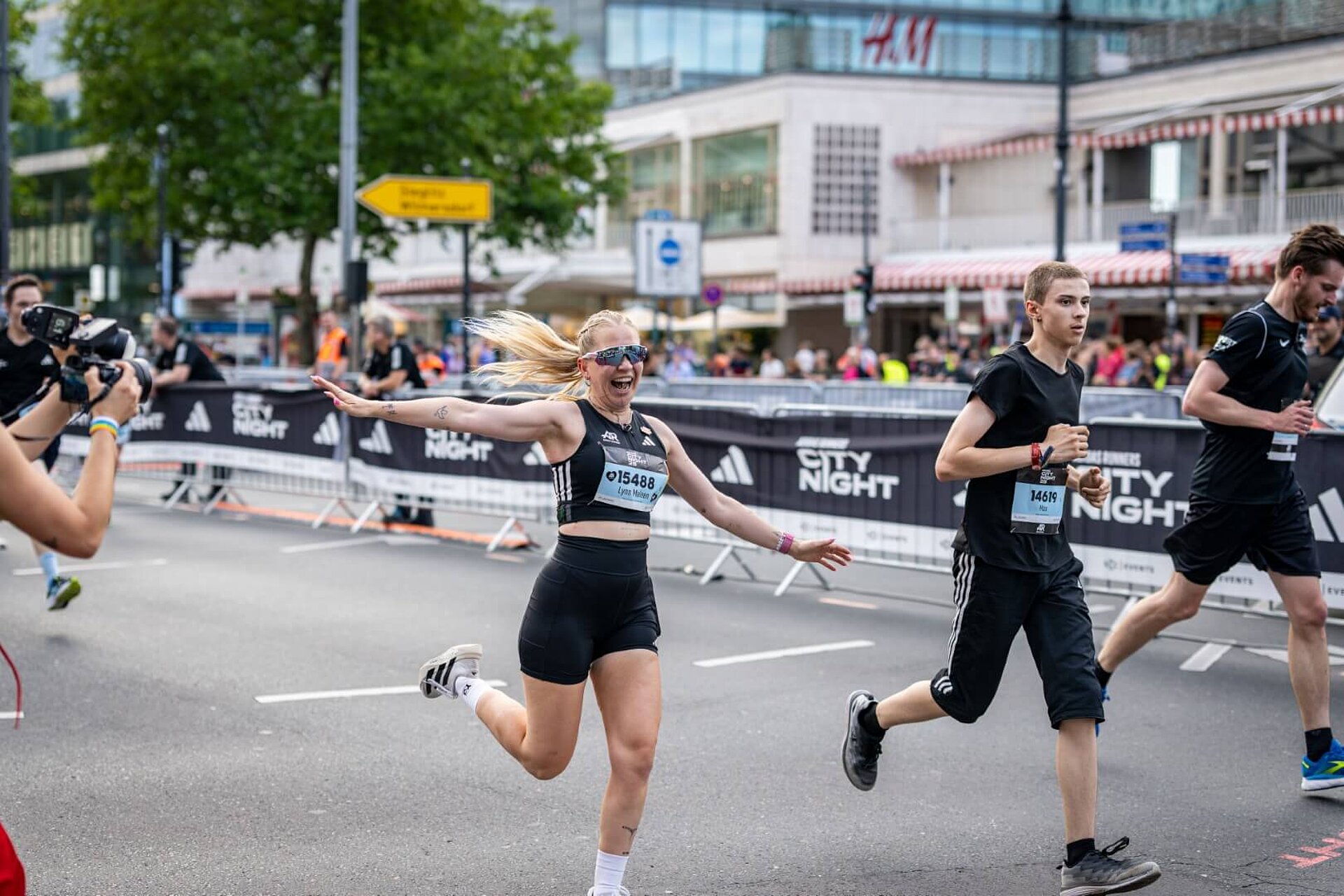 A runner celebrates with her arms outstretched. Two other runners are jogging beside her.