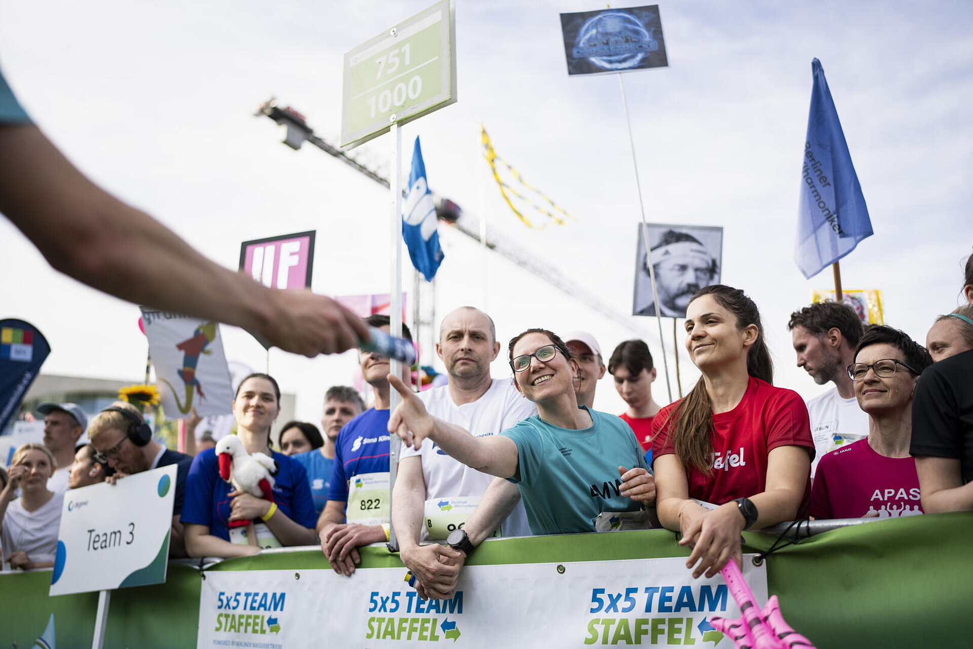 Lächelnde Teilnehmerin übernimmt Staffelstab. Um sie herum zahlreiche weitere Läufer:innen. Kreative Teamschilder und Fahnen ragen in die Luft.