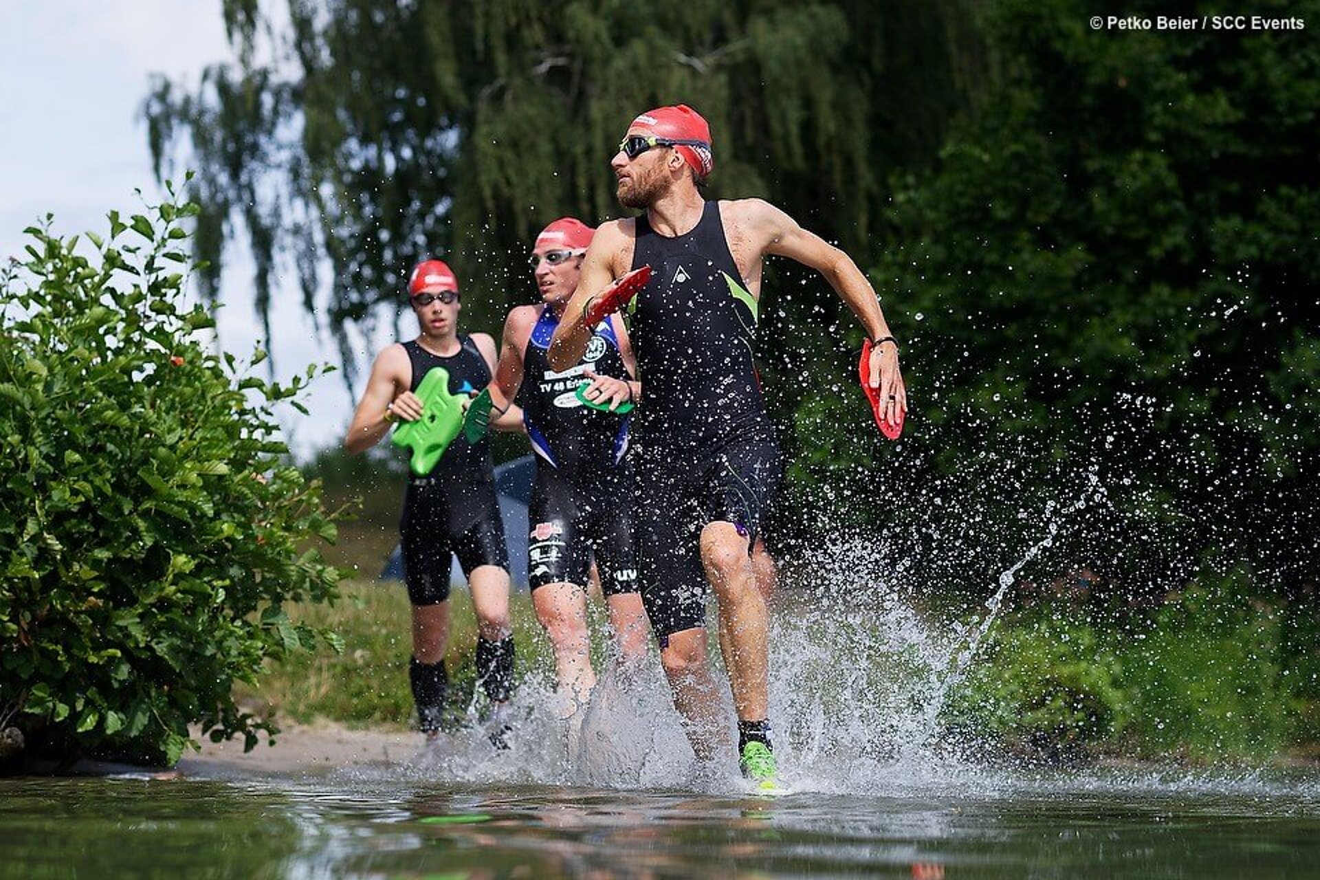 Faris Al-Sultan rennt spritzends mit Badekappe, Schwimmbrille und Paddles durchs Wasser. Hinter ihm folgen zwei weitere Teilnehmer.